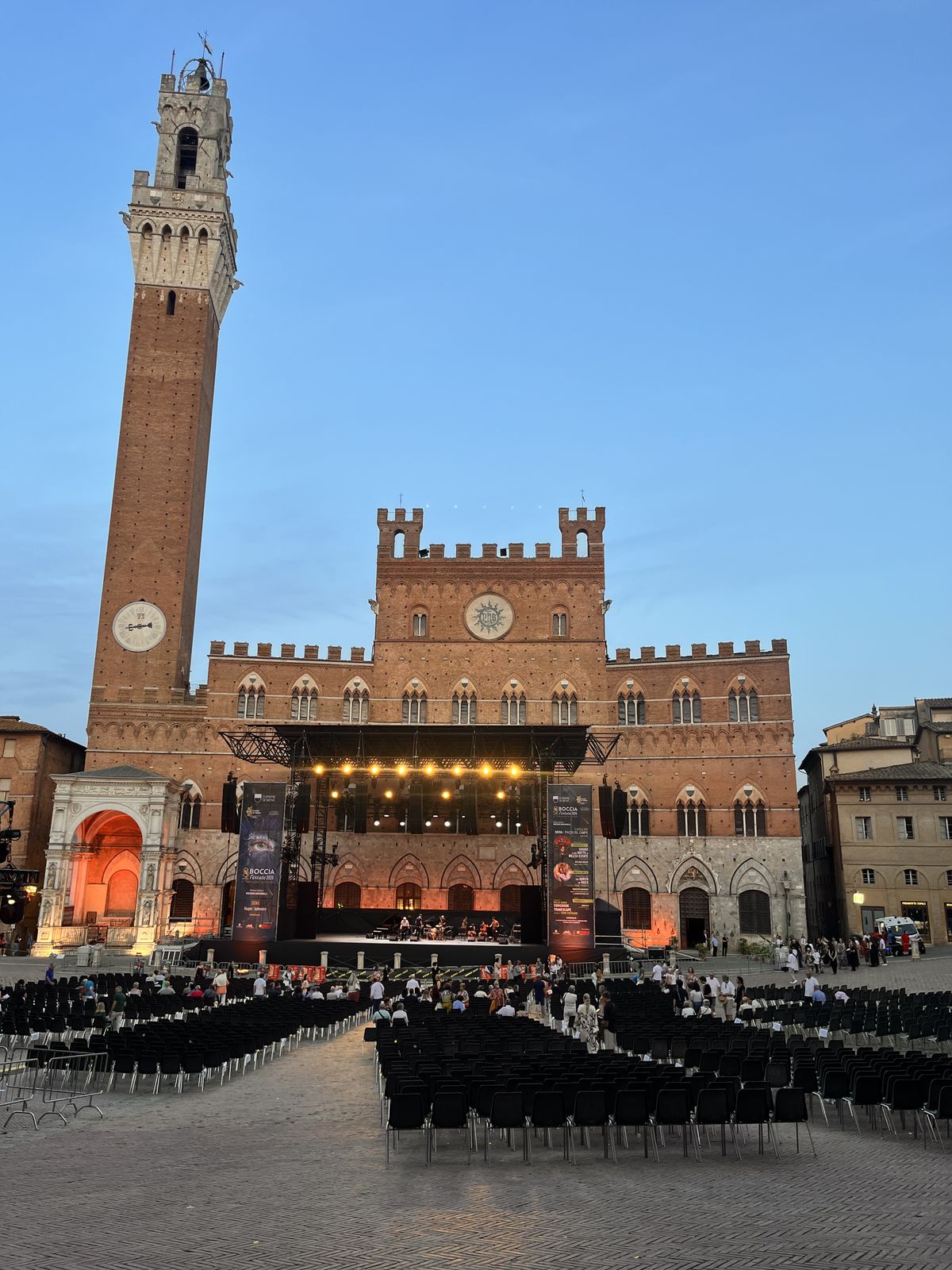 Concert stage set against the Palazzo Pubblico in Piazza del Campo at dusk, Torre del Mangia above, chair rows filling for Siena Jazz's free concert on 23 July 2026